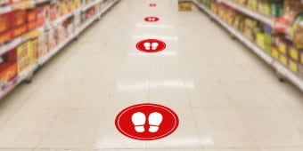 Floor graphics with white footprints in red circles show recommended social distancing in a grocery store aisle.
