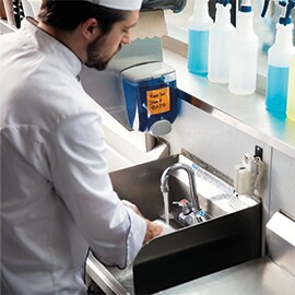 A chef washes his hands at a sink in a commercial kitchen with cleaning supplies nearby.