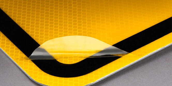 A closeup of a protective overlay film peeled back on top of a reflective yellow sign.
