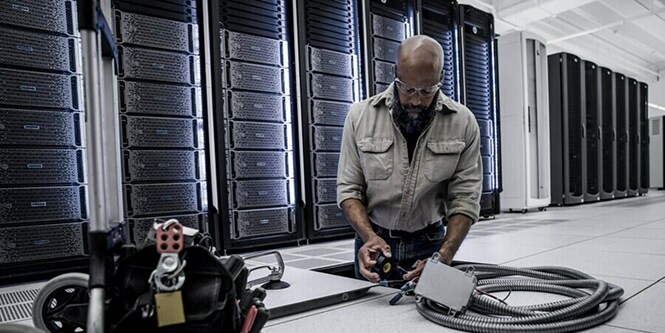 Técnico arrodillado en un centro de datos, trabajando con cables y herramientas frente a varias filas de racks de servidores.
