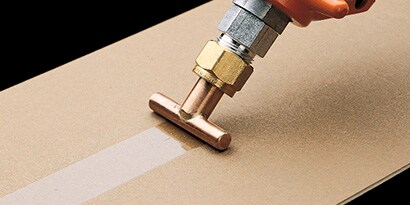 Image of a hot melt applicator with a spreader tip applying an even-width band of hot melt adhesive
