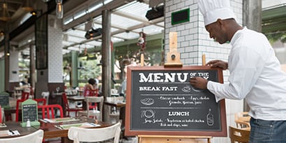 Un chef actualizando el tablero de menú de tiza que se muestra afuera de un restaurante comercial.

