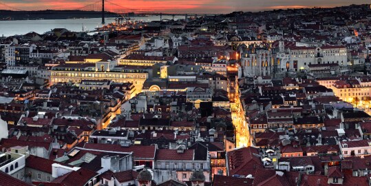 Vista panorámica de Lisboa al atardecer, con tejados rojizos en primer plano, calles iluminadas, edificios históricos en el centro y el río Tajo al fondo con un puente colgante bajo un cielo anaranjado.