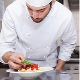 Un chef añade cuidadosamente un detalle a un plato de pasta en una cocina comercial.