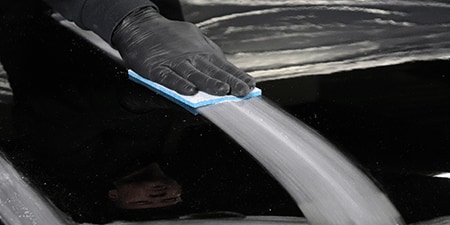 Una persona con guantes negros utiliza una almohadilla abrasiva azul para lijar una superficie negra brillante y crea una franja lisa mate.
