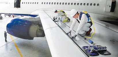 Workers on aircraft wing performing maintenance secured by fall protection gear
