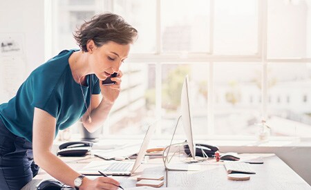 Mujer de pie en un escritorio, tomando notas y hablando por teléfono en una oficina iluminada por la brillante luz del sol a través de grandes ventanales.
