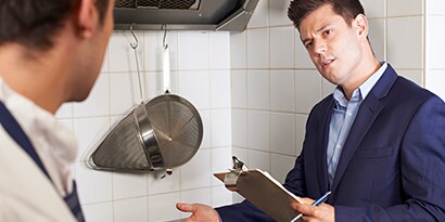Un hombre, vistiendo un traje y sosteniendo una tabla de apuntes, hace un gesto hacia una estufa de cocina comercial mientras un trabajador de cocina observa.
