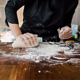 A cook presses dough on a floury wooden countertop, with a jar of flour nearby.
