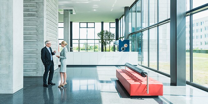 Interior de un edificio de oficinas moderno rodeado por paredes de ventanas con dos profesionales de negocios conversando.
