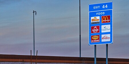 A blue exit sign listing restaurants in front of a dusk sky.
