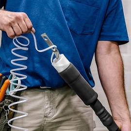 Trabajador con camisa azul sosteniendo una manguera de aire enrollada conectada a una herramienta neumática negra, con cinturón de herramientas y pantalones caqui visibles.